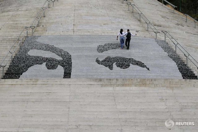 Una pareja sube un tramo de escaleras con azulejos que representan los ojos del fallecido presidente de Venezuela Hugo Chávez, en Caracas, 5 de marzo de 2015.  En los últimos años, la imagen más omnipresente en Venezuela ha sido un esténcil de los ojos de Chávez mirando, paternalmente, desde vallas publicitarias, camisetas y viviendas de interés social. Con un simple cambio en la posición de las cejas y una ampliación de los ojos, sin embargo, los partidarios de la oposición han creado un meme que muestra un Chávez sorprendido por la primera derrota parlamentaria de sus partidarios.REUTERS/Jorge Silva