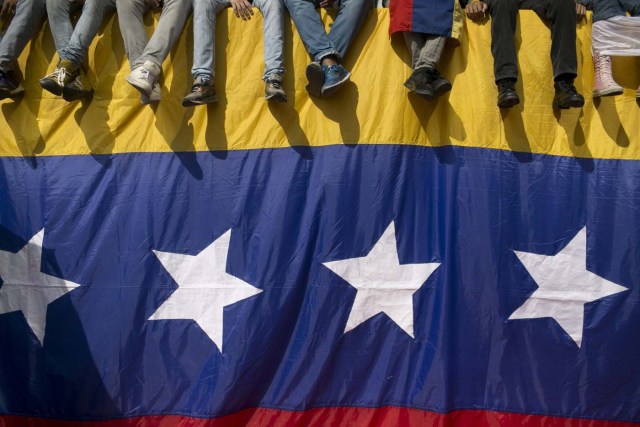 En esta imagen del 26 de octubre de 2016, gente sentada en un muro cubierto por una bandera venezolana durante una protesta contra el presidente Nicolás Maduro en Caracas, Venezuela. La tensión se agravó en Venezuela después de que la Asamblea Nacional votara a favor de abrir un juicio político contra Maduro por romper el orden constitucional, y de que los líderes opositores convocaran marchas masivas el miércoles para expulsar al mandatario. (AP Foto/Alejandro Cegarra)