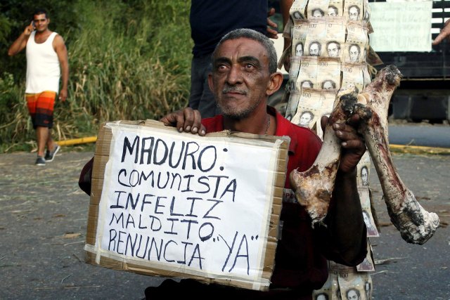 A man holds a bone and a placard that reads "Maduro: Communist, unhappy, damn. Resigns, now", in front of a pole covered with 100-bolivar bills during a protest in El Pinal