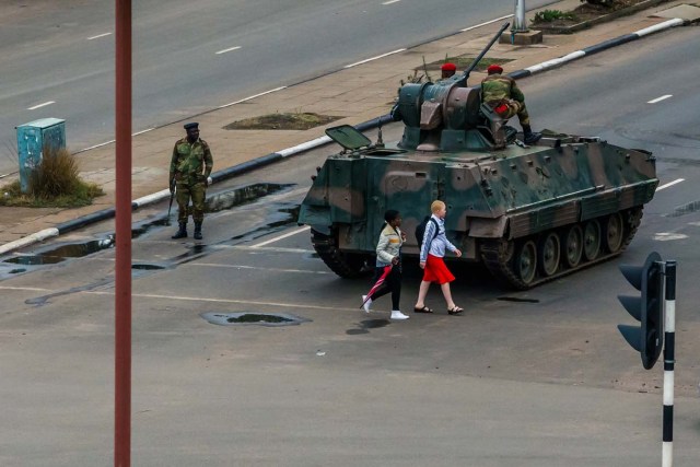 Young women walk past an armoured personnel carrier that stations by an intersection as Zimbabwean soldiers regulate traffic in Harare on November 15, 2017. Zimbabwe's military appeared to be in control of the country on November 15 as generals denied staging a coup but used state television to vow to target "criminals" close to President Mugabe. / AFP PHOTO / -